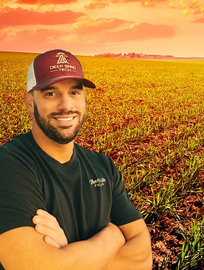 Man in cap and black shirt, arms crossed, smiling in a field with a sunset background.